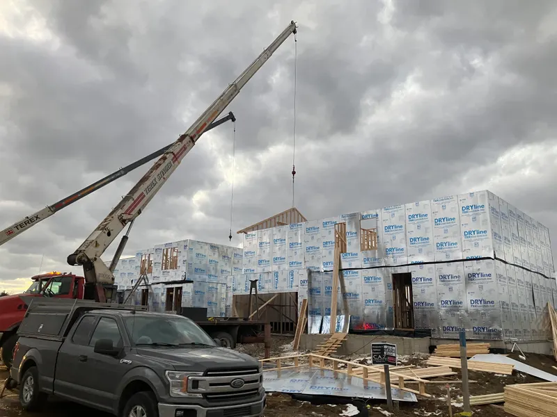 A ROC Crane boom truck lifting a framed wall panel into place on a two-storey building under construction.